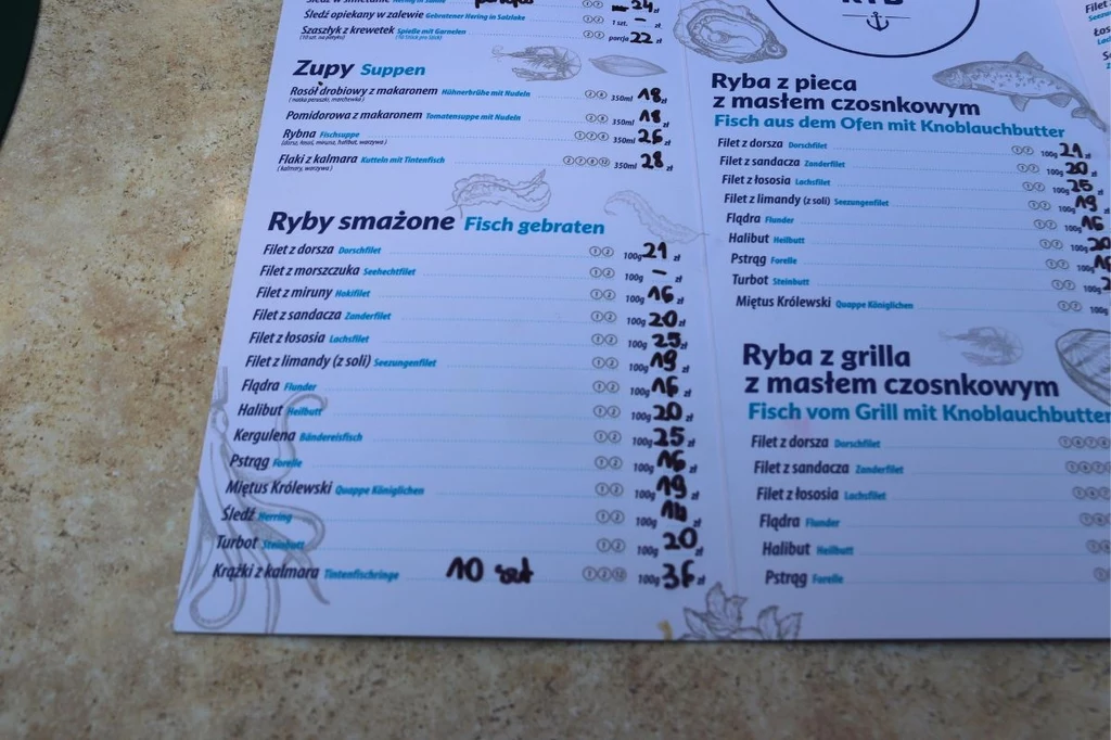 W smażalniach waga jednej porcji ryby to najczęściej 200-400 g Menu restauracyjne z sekcjami dotyczącymi zup oraz dań rybnych smażonych, z pieca i z grilla z masłem czosnkowym, wraz z widocznymi cenami obok każdego dania, tekst w języku polskim i niemieckim.