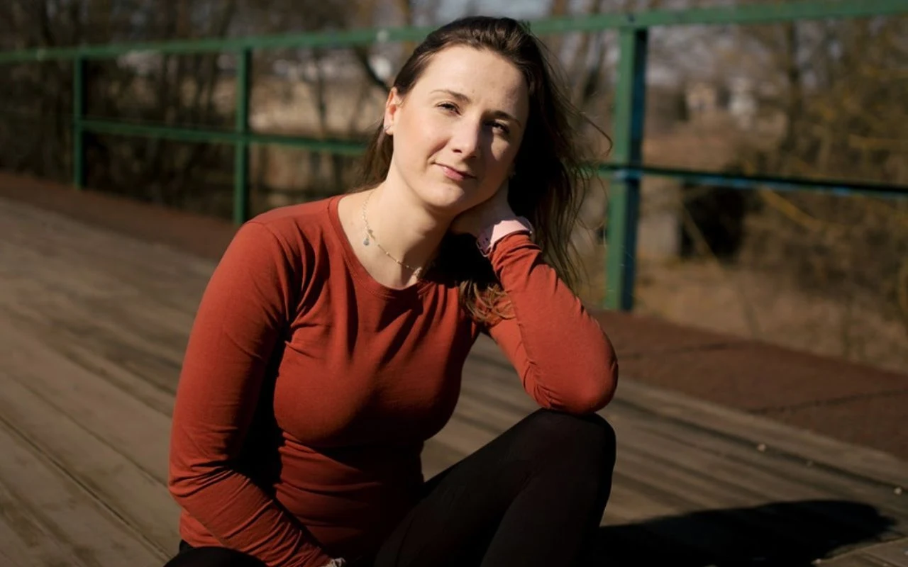 Młoda kobieta w bordowej bluzce siedzi na drewnianym podeście na zewnątrz, opierając rękę o głowę, w tle metalowa barierka i drzewa.