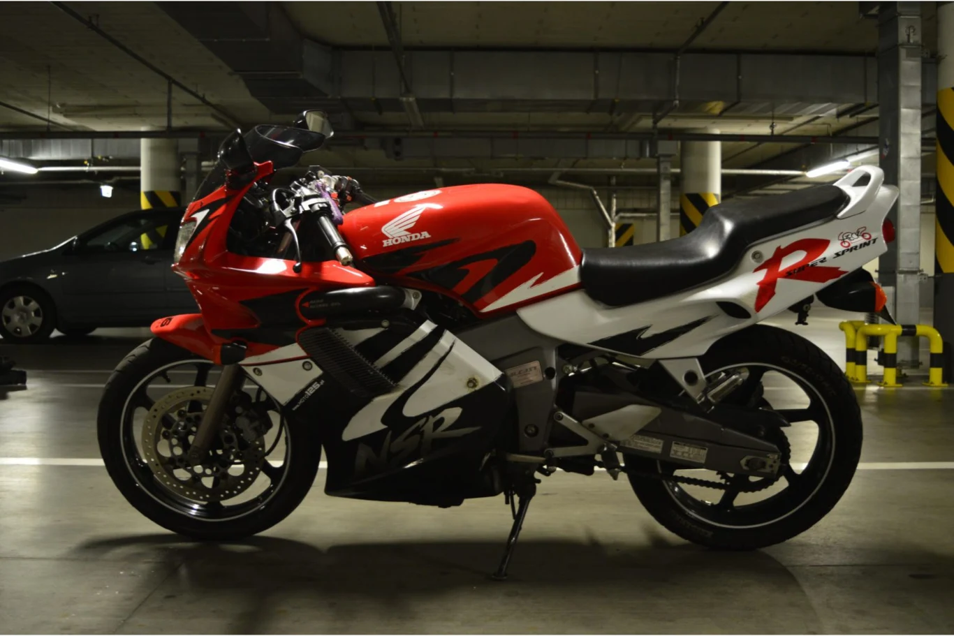 Czerwony sportowy motocykl Honda zaparkowany w podziemnym garażu, w tle widać szare auto oraz betonowe filary i żółto-czarne oznakowania zabezpieczające narożniki.