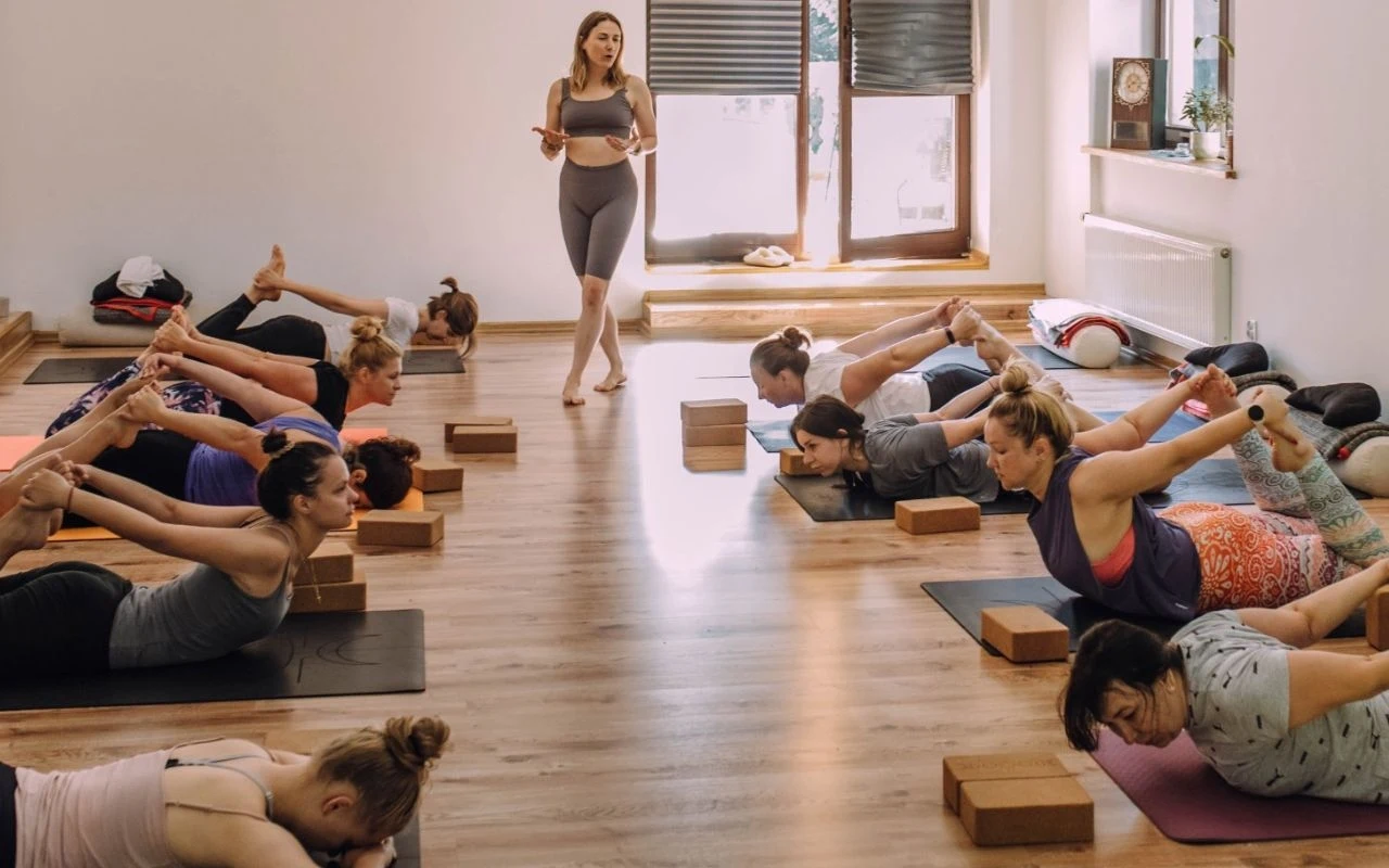 Grupa osób wykonuje ćwiczenia jogi na matach pod okiem instruktorki, korzystając z drewnianych klocków i pasów do jogi w jasnej, przestronnej sali z dużymi oknami.