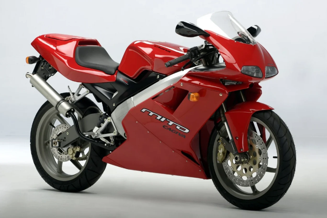 Cagiva Mito 125 to najmocniejszy, najszybszy i najbardziej zaawansowany motocykl o pojemności 125 cm3, który podbił rynek w latach 90. Czerwony sportowy motocykl z aerodynamiczną owiewką, srebrnym układem wydechowym oraz srebrno-czarnymi detalami na tle jasnego, neutralnego tła.