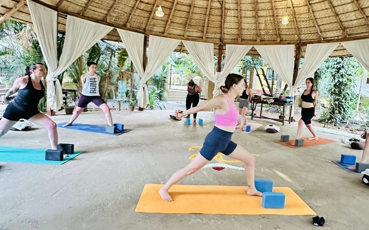 Grupa osób ćwicząca jogę w przestronnej altanie z dachem krytym strzechą, w otoczeniu naturalnej zieleni, każdy uczestnik używa maty oraz kostek do jogi, zapewniając komfortowe warunki do ćwiczeń.