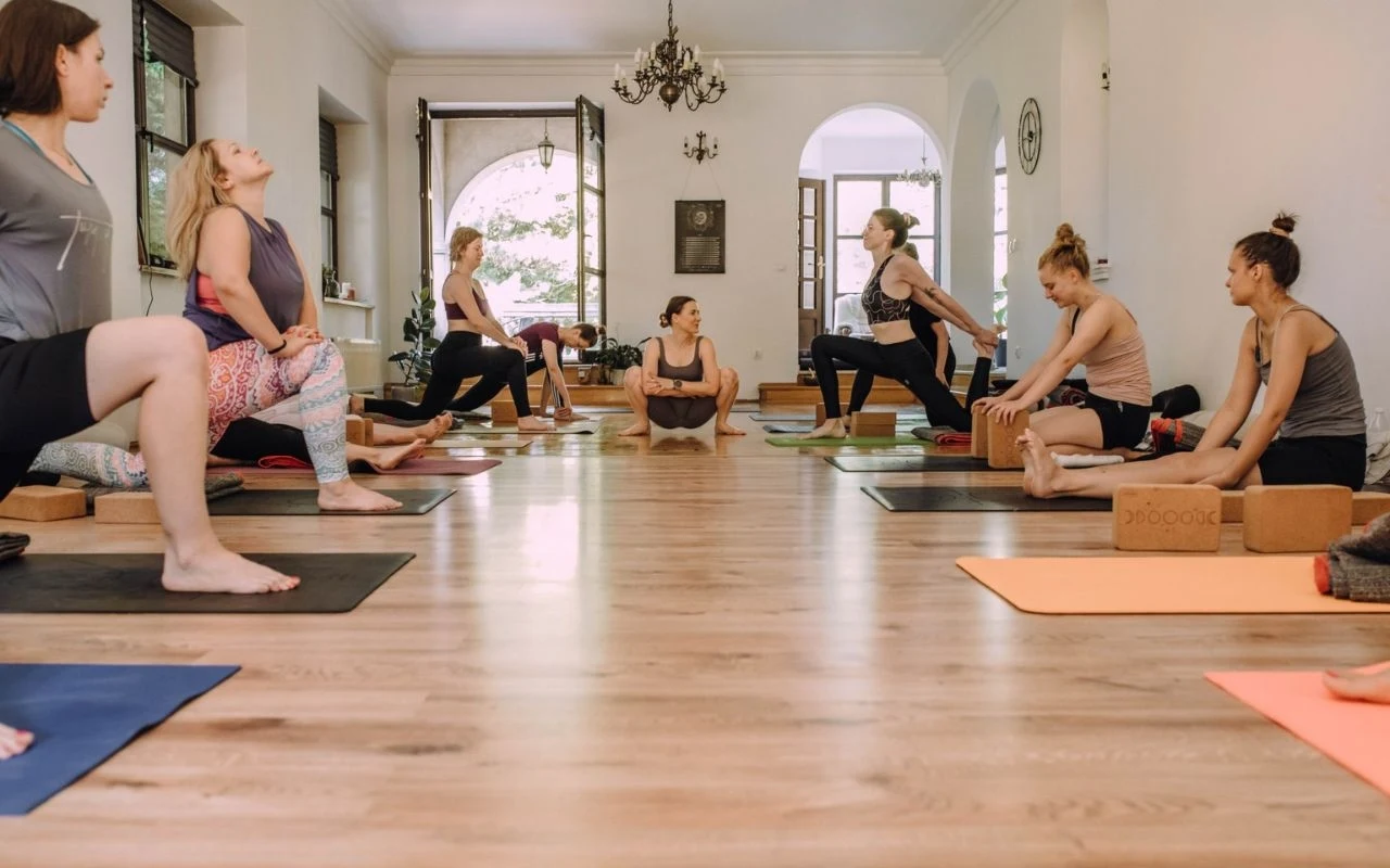 Grupa kobiet ćwicząca jogę na matach w jasnym, przestronnym pomieszczeniu ze stylowymi żyrandolami, dużymi oknami i drewnianą podłogą, uczestniczki wykonują pozycje rozciągające prowadzone przez instruktorkę.