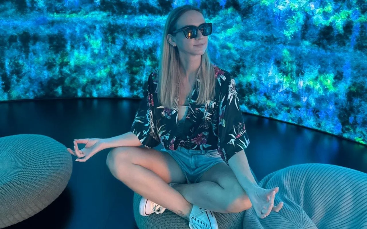 Młoda kobieta w okularach przeciwsłonecznych siedzi po turecku na poduszce, gestykulując jak podczas medytacji, w tle podświetlona, abstrakcyjna ściana w odcieniach niebieskiego i zielonego, atmosfera relaksu i wyciszenia.