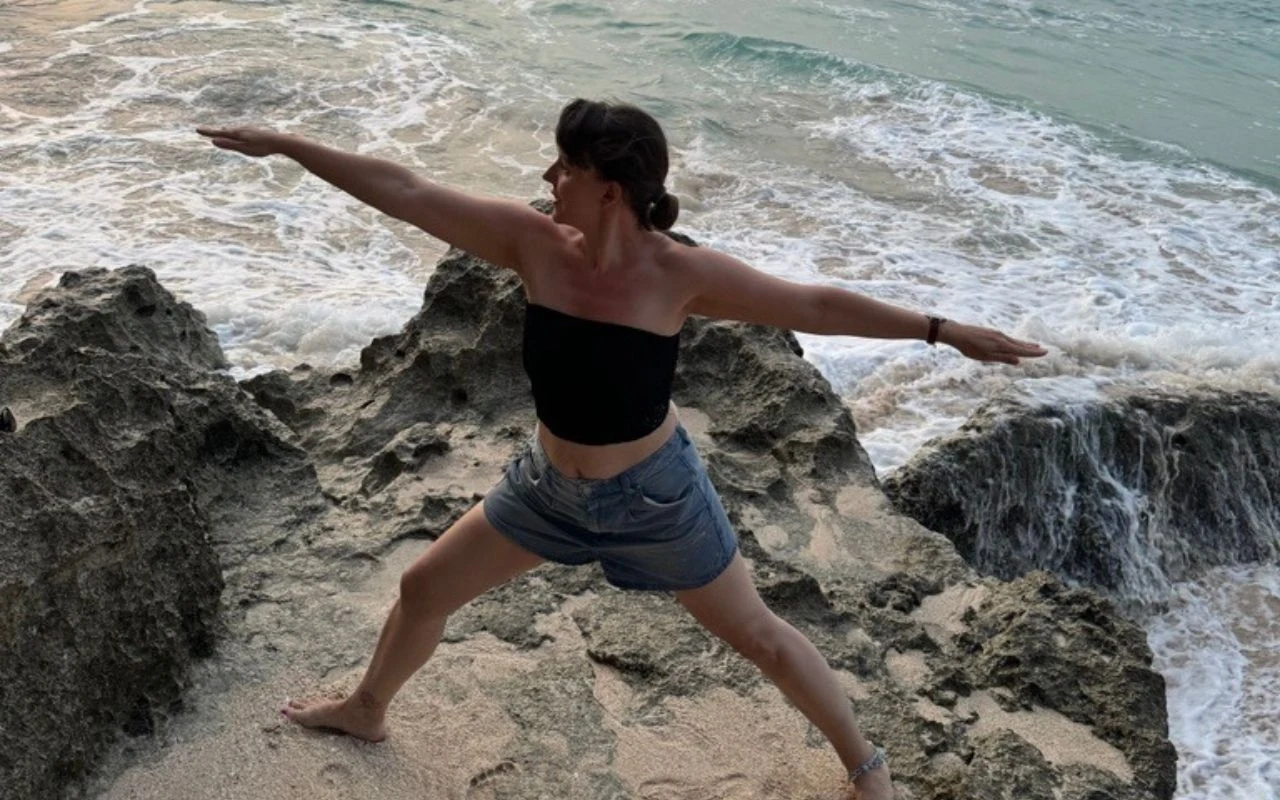 Kobieta wykonująca pozycję jogi na skalistym brzegu przy falującym morzu, ubrana w czarny top i szorty, otoczona naturalnym krajobrazem.