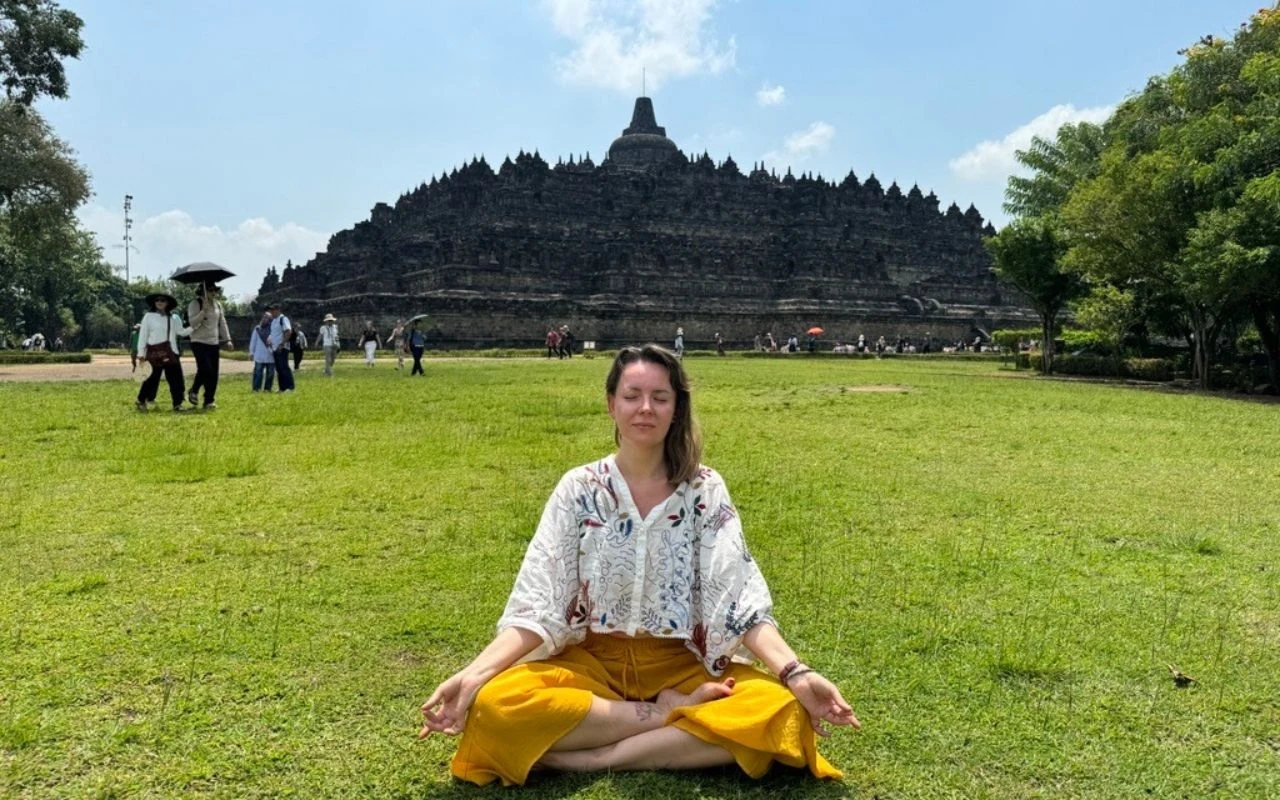 Kobieta siedzi w pozycji medytacyjnej na trawie, w tle monumentalna świątynia Borobudur oraz kilka osób spacerujących po zielonym terenie.