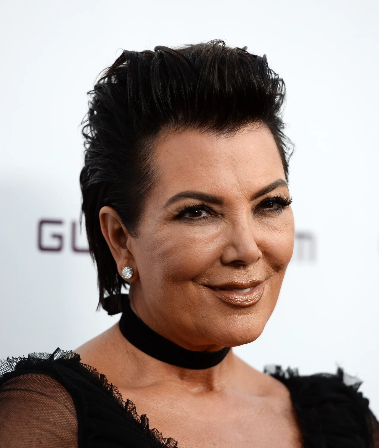 Jeszcze do niedawna twarz Kris Jenner wyglądała tak Uśmiechnięta kobieta z krótkimi, ciemnymi włosami i perfekcyjnym makijażem nosi czarną suknię z tiulowymi elementami i elegancki czarny choker, ozdobiona kolczykami w kształcie okrągłych kryształów, na jasnym tle z nieostrym logotypem.