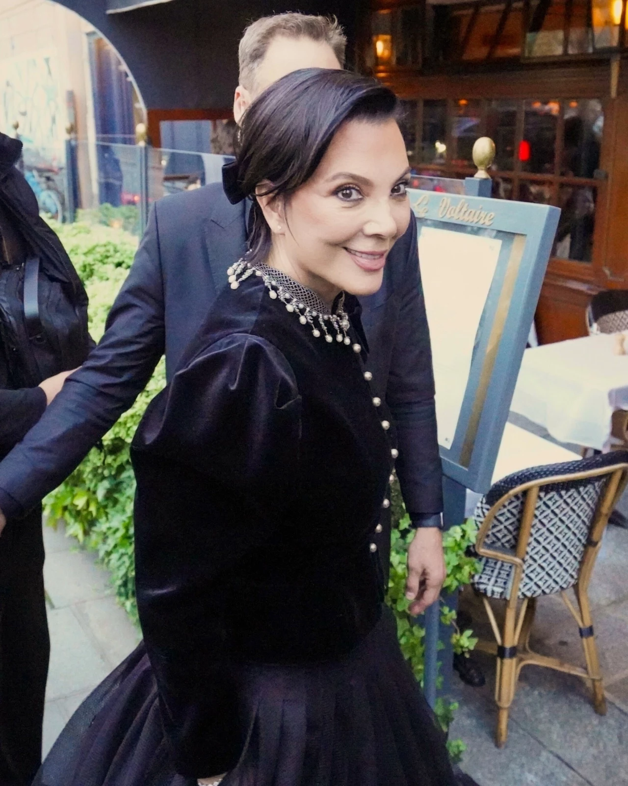 Nowy wygląd Kris Jenner zaskakuje fanów Elegancko ubrana kobieta w czarnej kreacji z perłowym naszyjnikiem stoi uśmiechając się na tle restauracyjnego ogródka; w tle stolik z krzesłami, tablica restauracyjna oraz elementy roślinności.