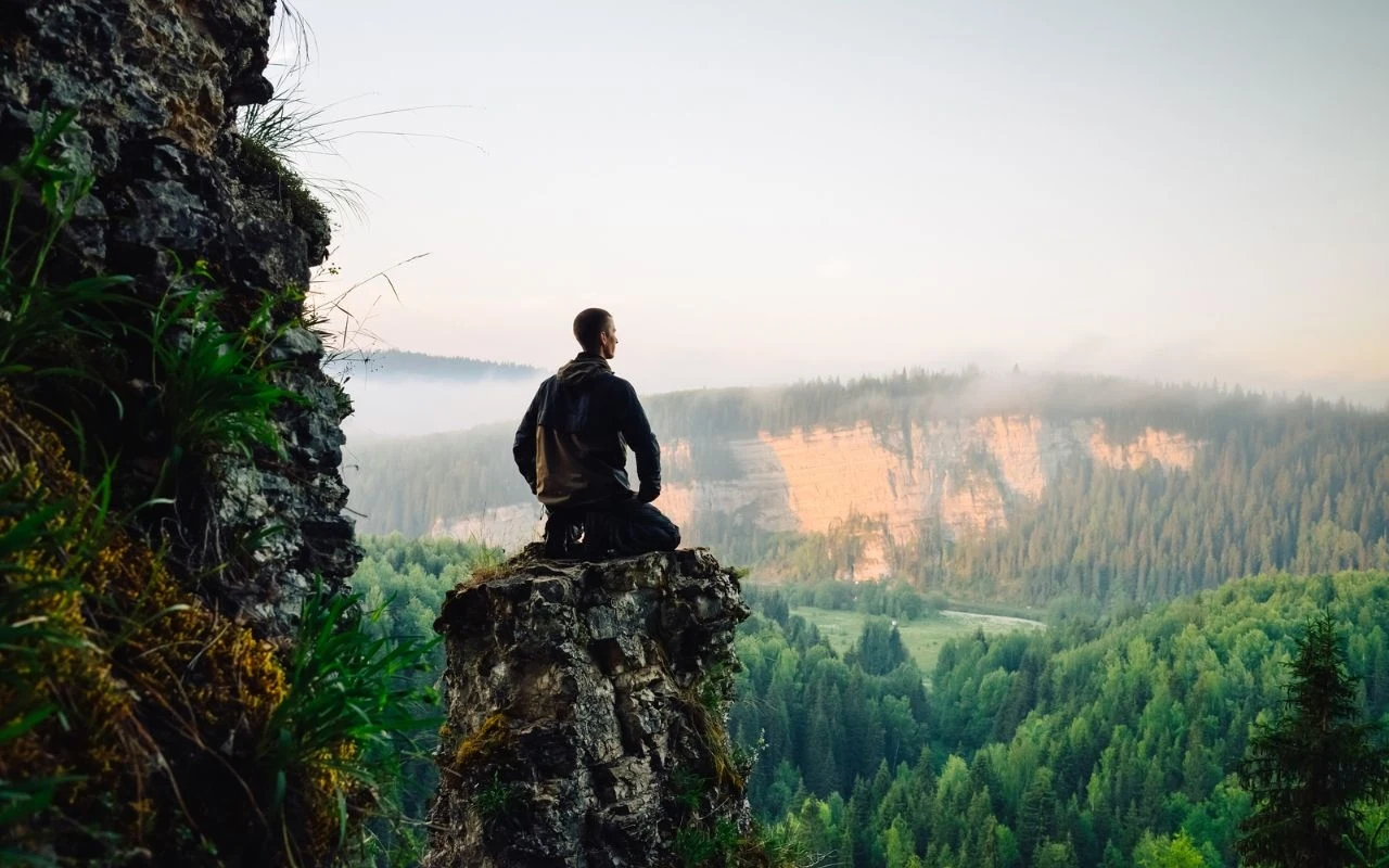 Mężczyzna siedzi na krawędzi skalnego urwiska, otoczony dziką roślinnością, spoglądając na rozległą panoramę leśnych wzgórz, nad którymi unosi się delikatna mgła i zachodzące światło słoneczne.