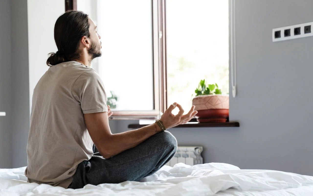 Mężczyzna siedzi po turecku na białym łóżku, zwrócony w stronę okna, wykonując gest medytacji, w tle widoczna doniczka z zieloną rośliną.
