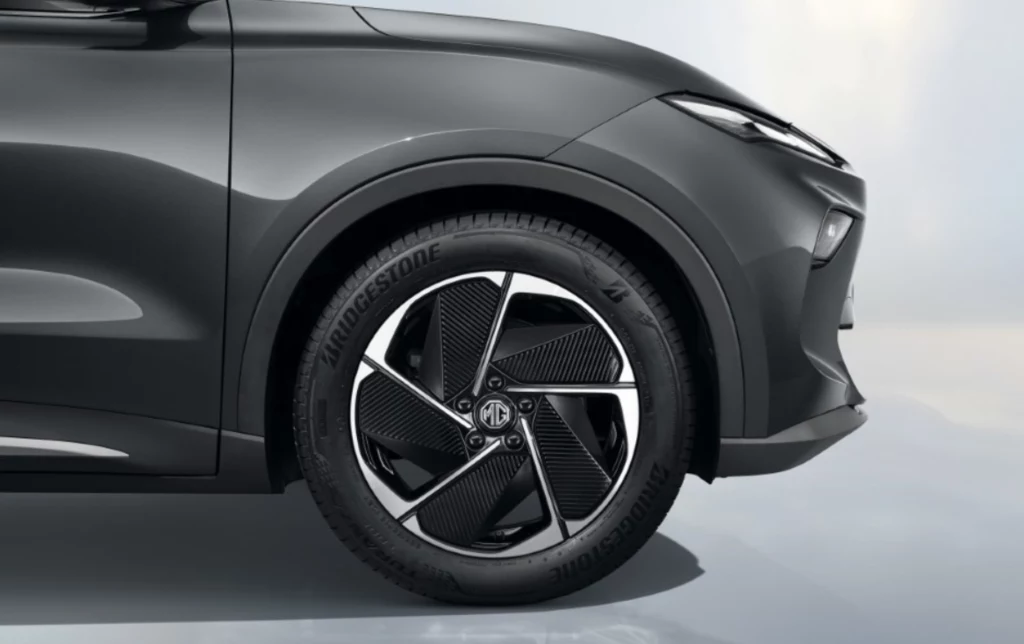 MGS5 EV uzyskał najwyższą ocenę 5 gwiazdek w testach Euro NCAP. Czarna felga z charakterystycznym wzorem, opona marki Bridgestone oraz fragment przedniego błotnika i reflektora nowoczesnego samochodu osobowego w szarym kolorze.