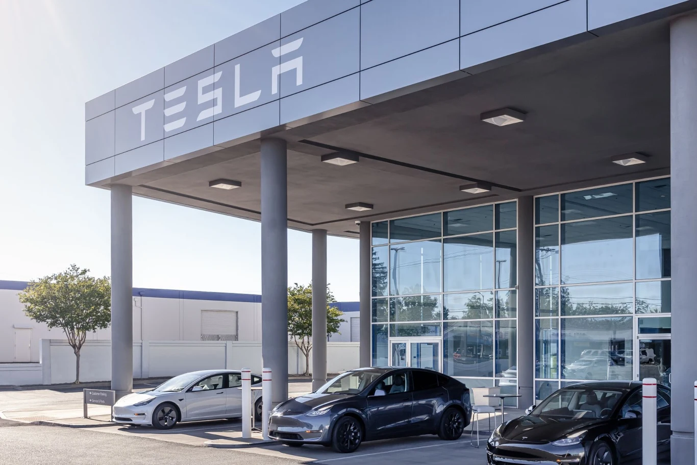 Tesla Center Wrocław otwiera się już w czwartek. Salon samochodowy Tesla z trzema nowoczesnymi samochodami elektrycznymi zaparkowanymi przed szklaną fasadą budynku, logo firmy widoczne nad wejściem, w tle jasna zabudowa i kilka drzew.