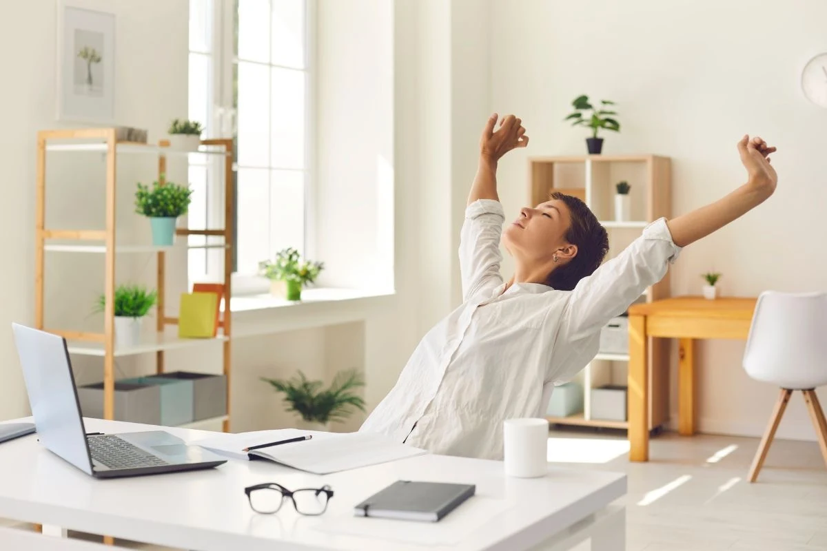 Balans pomiędzy pracą a czasem wolnym jest kluczowy dla Elastyczne zarządzanie czasem pracy pozwala lepiej reagować na potrzeby zawodowe i prywatne Kobieta siedząca przy biurku w jasnym, nowoczesnym biurze, z rozciągniętymi do góry ramionami, wyrażająca odprężenie; na biurku znajdują się laptop, notatnik, okulary i kubek, a w tle rośliny oraz regały z segregatorami.