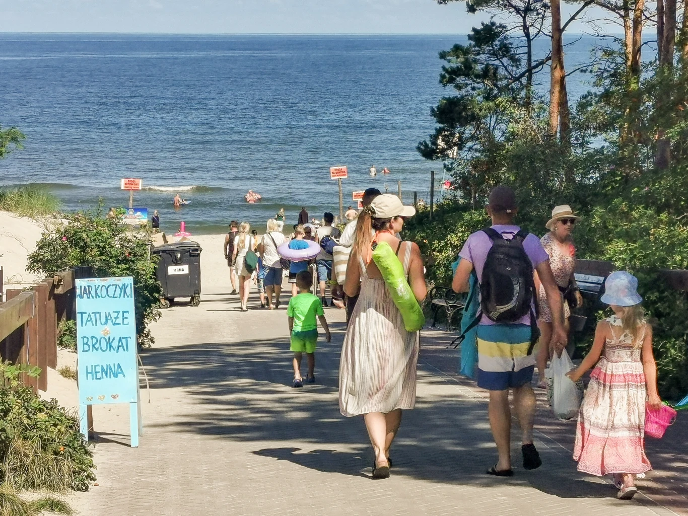 Nowe pamiątki opanowały stoiska nad Bałtykiem Grupa ludzi spaceruje deptakiem prowadzącym do plaży, część osób niesie ręczniki i parasole, po bokach roślinność, w tle Morze Bałtyckie oraz kąpiący się i spacerujący ludzie, widoczna tablica z ofertą usług wakacyjnych.