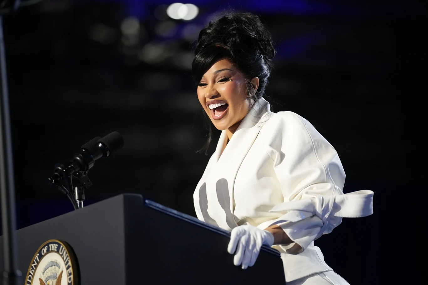 Cardi B ogłasza premierę drugiego albumu Kobieta ubrana na biało stoi za mównicą z mikrofonem, uśmiecha się szeroko i sprawia wrażenie radosnej podczas publicznego przemówienia.