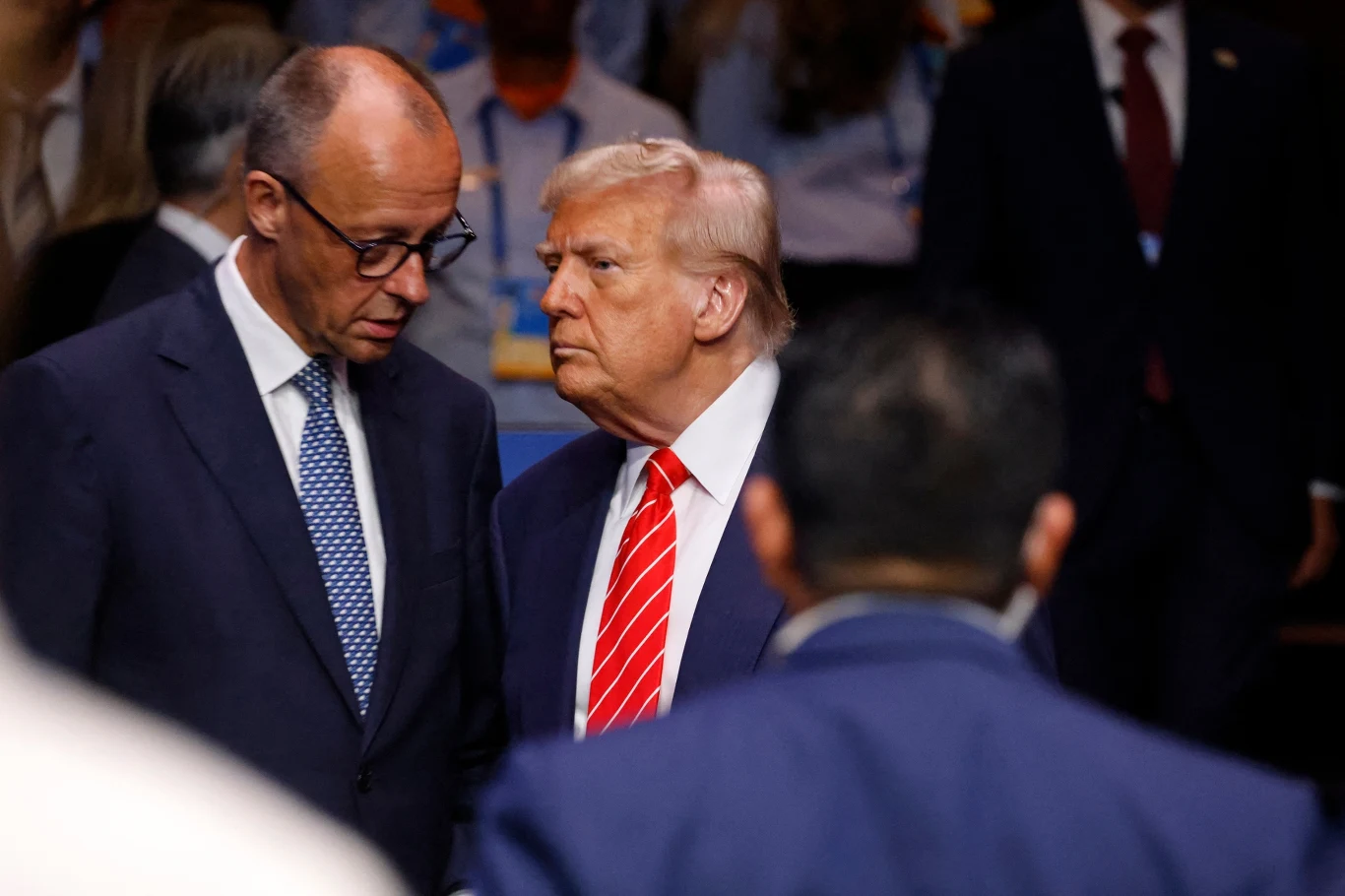 Dwóch mężczyzn w garniturach prowadzi poważną rozmowę w otoczeniu innych osób, jeden z nich ma jasne włosy i czerwony krawat, drugi okulary i niebieski krawat w kropki.