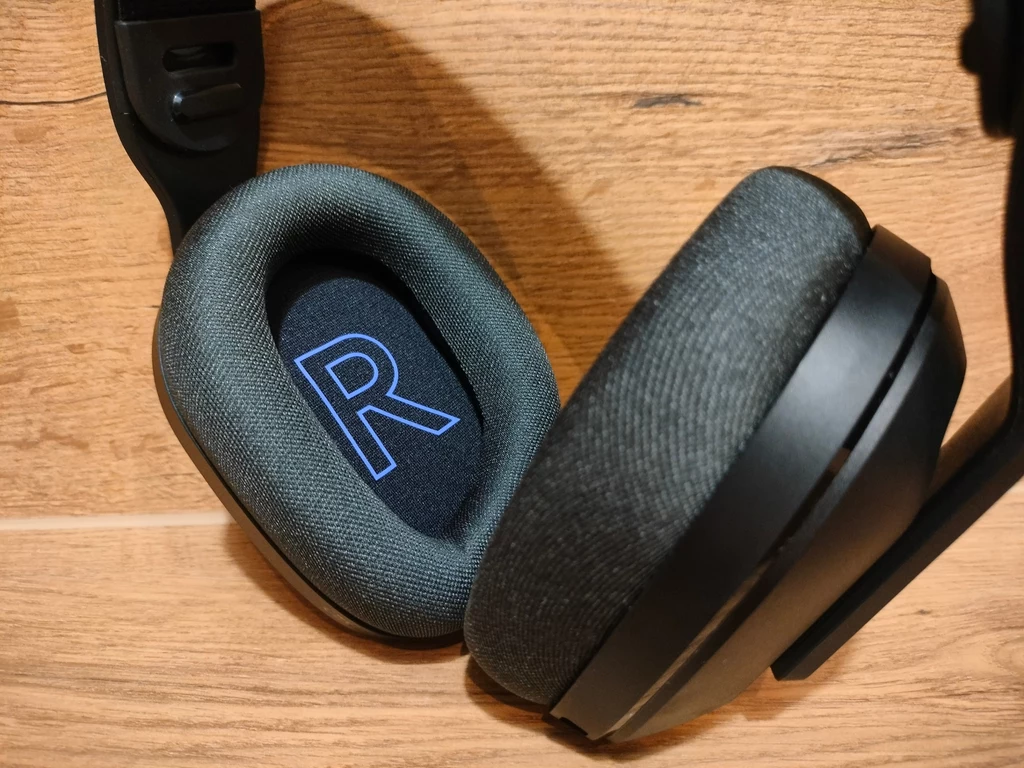 Logitech G522 Lightspeed Czarne nauszne słuchawki z miękkimi poduszkami, leżące na jasnym drewnianym tle, z widoczną literą R oznaczającą stronę słuchawek.