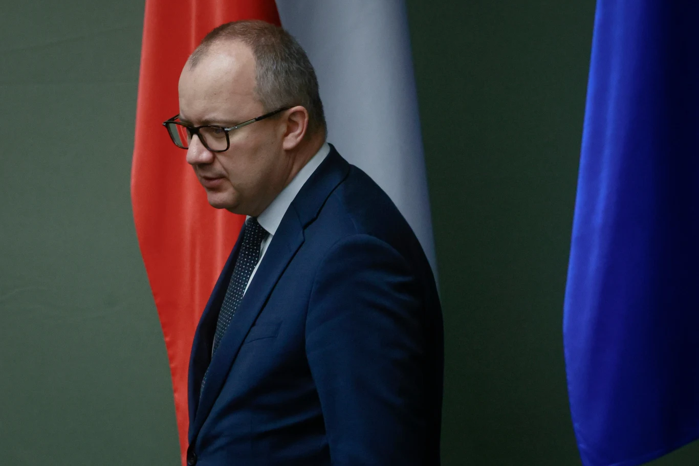 Prokurator generalny i minister sprawiedliwości Adam Bodnar Mężczyzna w garniturze i okularach idzie na tle flagi Polski, białej tkaniny i niebieskiej flagi.