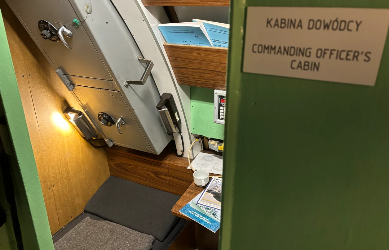 Wnętrze niewielkiej kabiny ze składanym łóżkiem, lampką, dokumentami oraz kubkiem na drewnianym blacie, tabliczka z napisem informującym o kabinie dowódcy po polsku i angielsku