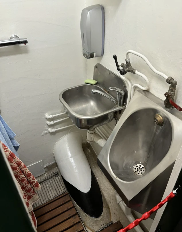 Mała łazienka o niestandardowej aranżacji, w której obok siebie zamontowano umywalkę, pisuar i fragment dużego pocisku lub torpedy. Wszystko wykonane z metalu, instalacje wodne są wyeksponowane, a na dozowniku mydła widać mydło w kostce.