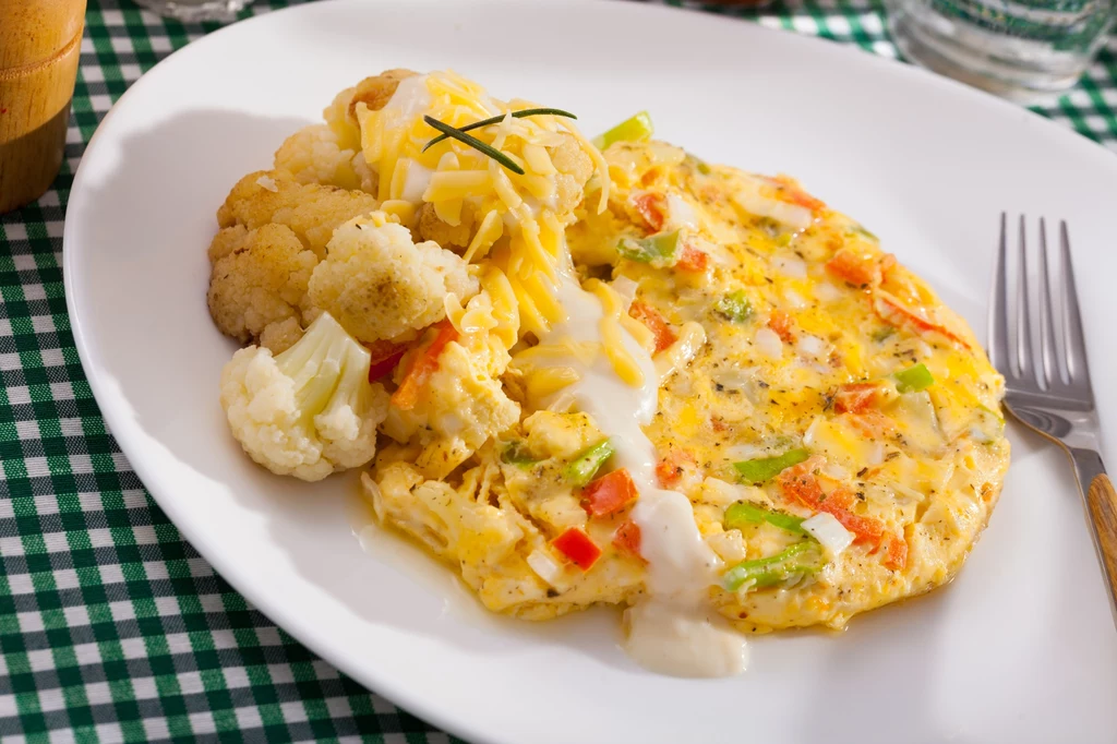 Omlet z warzywami, takimi jak papryka i cebula, polany sosem, podany z gotowanym kalafiorem, na białym talerzu ustawionym na zielono-białej kraciastej serwetce, obok widelec.