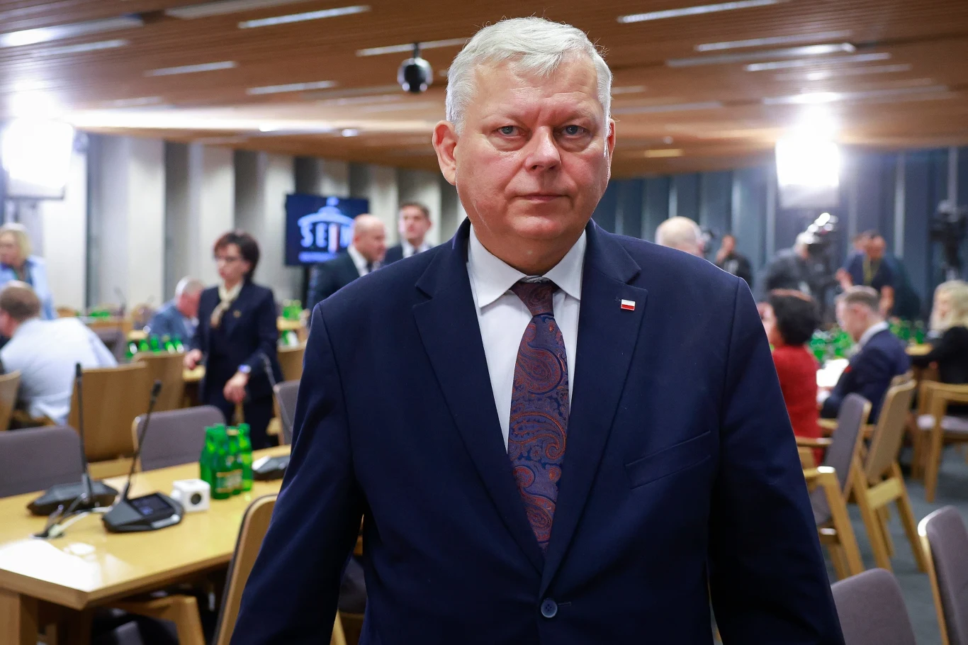 Poseł PiS Marek Suski przeszedł zawał serca Mężczyzna w garniturze z poważnym wyrazem twarzy stoi na pierwszym planie w sali konferencyjnej, w tle obecni są inni ludzie zajęci rozmowami i pracą przy stołach, na których stoją zielone butelki.
