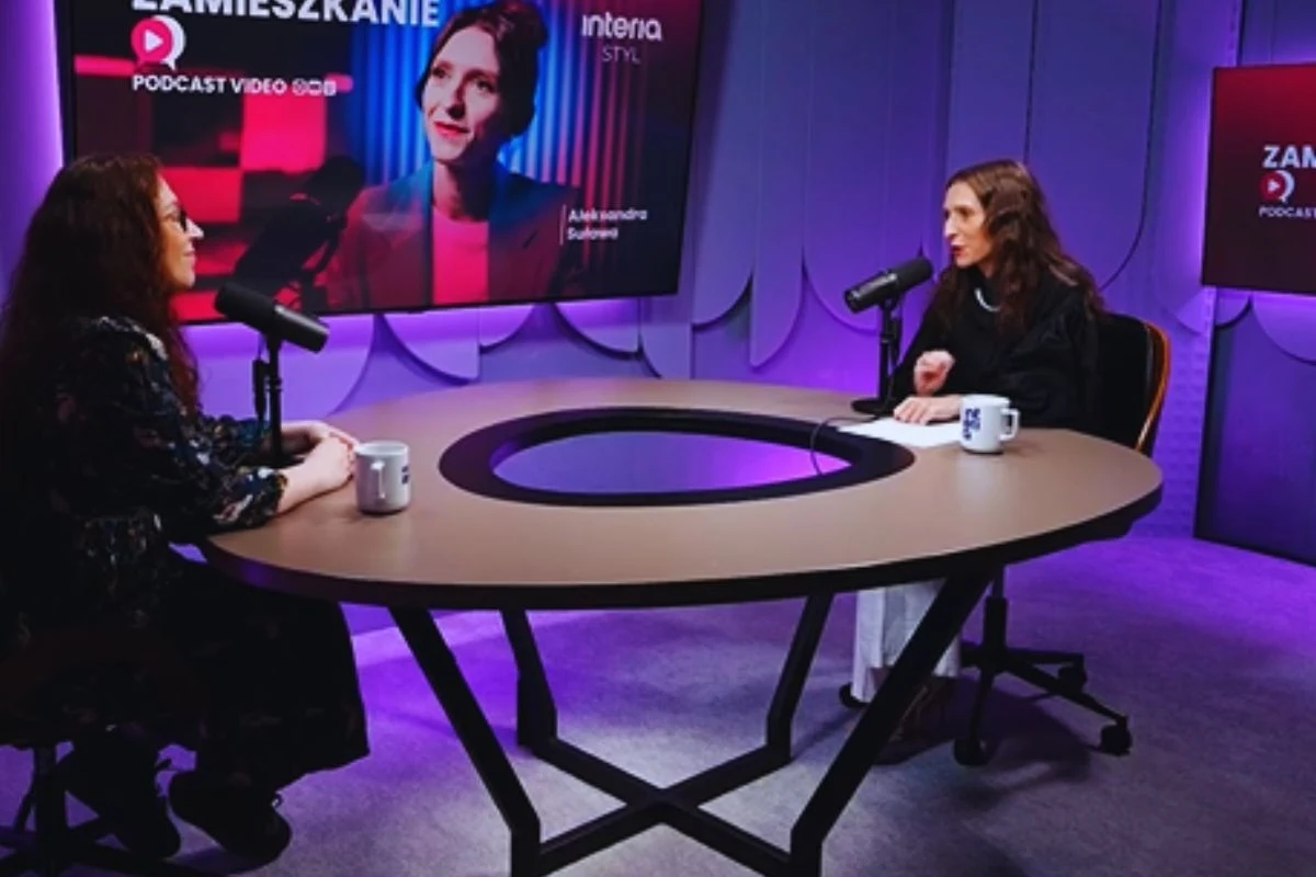 Dwie kobiety siedzą przy okrągłym stole, rozmawiają podczas nagrania podcastu, przed nimi znajdują się mikrofony i kubki. Na ścianie za nimi widoczny ekran z nazwą programu oraz portretem jednej z kobiet.