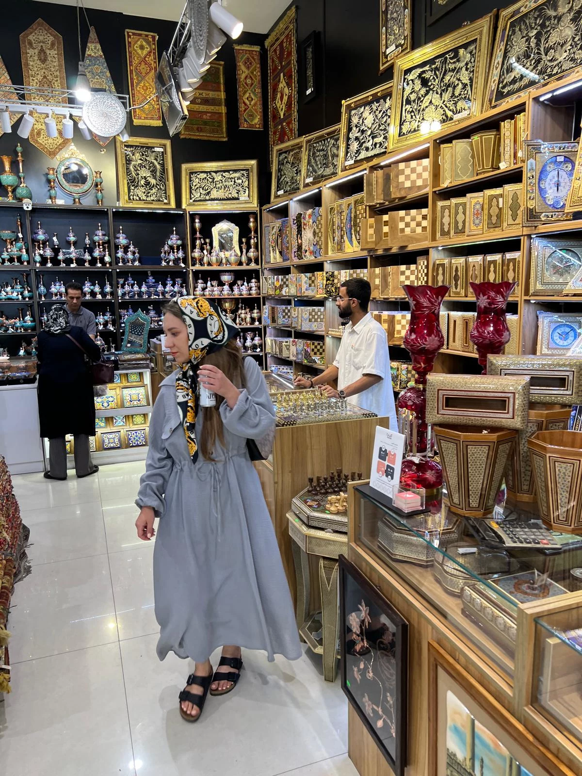 Iran Wnętrze sklepu z rękodziełem artystycznym, ściany i półki wypełnione bogato zdobionymi drewnianymi pudełkami, wazami i innymi wyrobami dekoracyjnymi, kobieta w jasnej sukni i z chustą na głowie ogląda ekspozycję, obsługiwany jest mężczyzna przy ladzie.