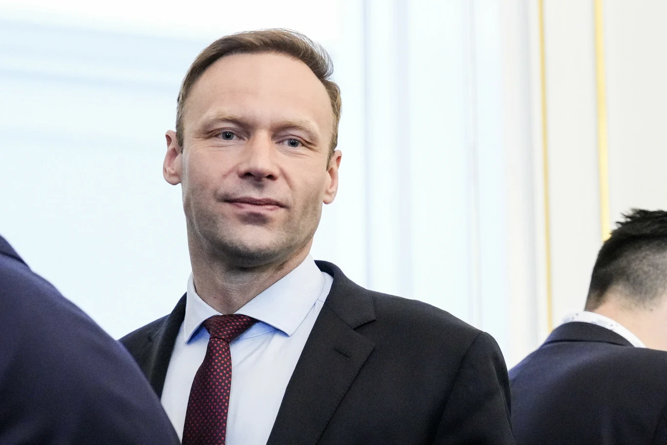 Mastalerek krytycznie o słowach Tuska Mężczyzna w garniturze i czerwonym krawacie stoi w jasnym pomieszczeniu, w tle widoczne sylwetki innych osób.