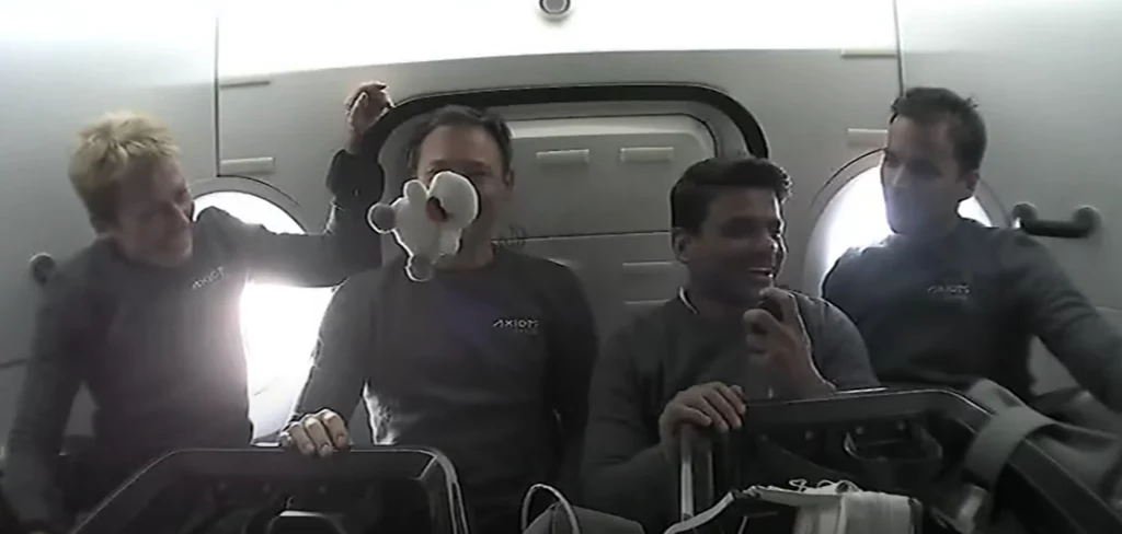 Po twarzach astronautów widać, że imię łabędzia nie było przypadkowe Cztery osoby w kombinezonach siedzą razem w jasnym wnętrzu, jedna z nich trzyma przed twarzą maskotkę unoszącą się w powietrzu, pozostali się uśmiechają, atmosfera jest radosna i swobodna.