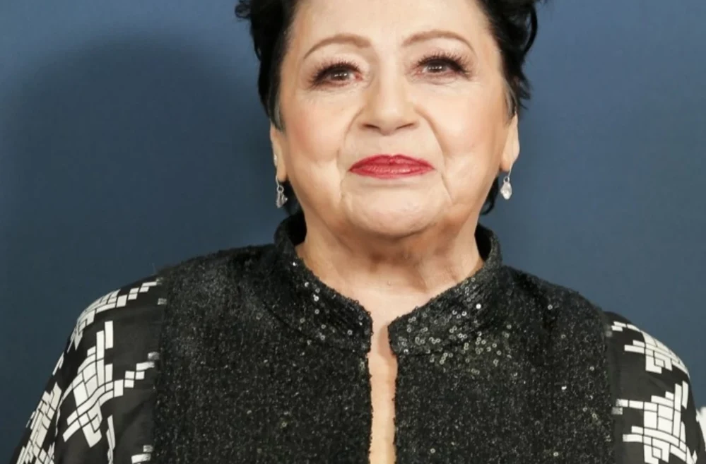 Ewa Bem opowiedziała o swoim stanie w ostatnim wywiadzie Starsza kobieta z uśmiechem na twarzy ubrana w ozdobną czarną marynarkę z błyszczącymi akcentami i geometrycznym wzorem na ramionach, na tle jednolitej, ciemnoniebieskiej ściany.