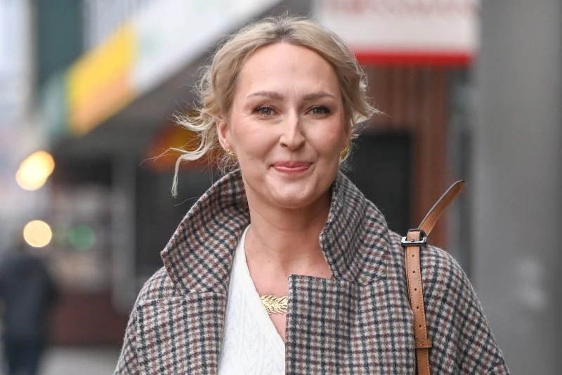 Anita Lipnicka Uśmiechnięta kobieta o blond włosach ubrana w wzorzystą kratę płaszcz i białą bluzkę, na ramieniu ma brązową torebkę, w tle rozmyte elementy miasta.