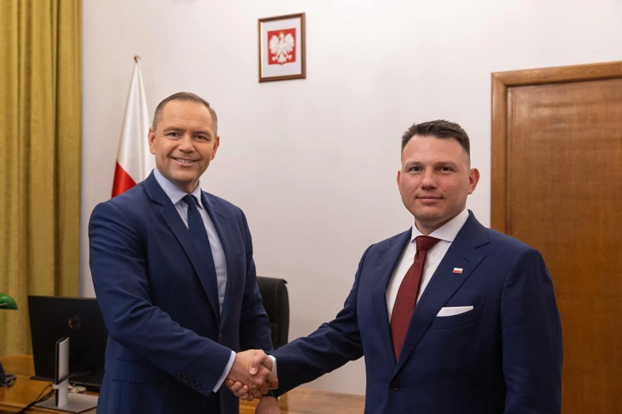 Sławomir Mentzen spotkał się z Karolem Nawrockim Dwóch mężczyzn w granatowych garniturach podaje sobie dłonie na tle biura, w którym znajduje się polska flaga oraz godło Polski na ścianie, atmosfera formalna, oficjalna.