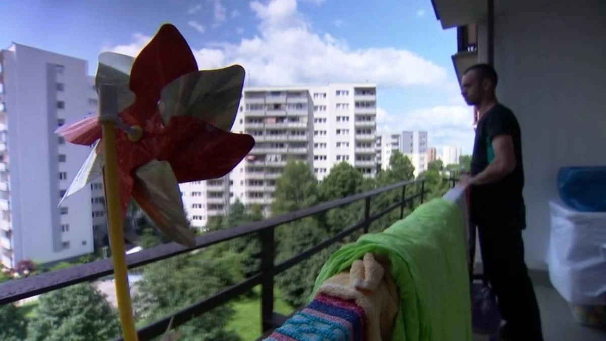 Śmierć czteromiesięcznego Oskara w rodzinie zastępczej. Biologiczna matka chłopca opuści więzienie Mężczyzna stojący na balkonie bloku mieszkalnego z suszącym się praniem oraz kolorowym wiatraczkiem na pierwszym planie, w tle widać osiedle wysokich budynków i zieleń.