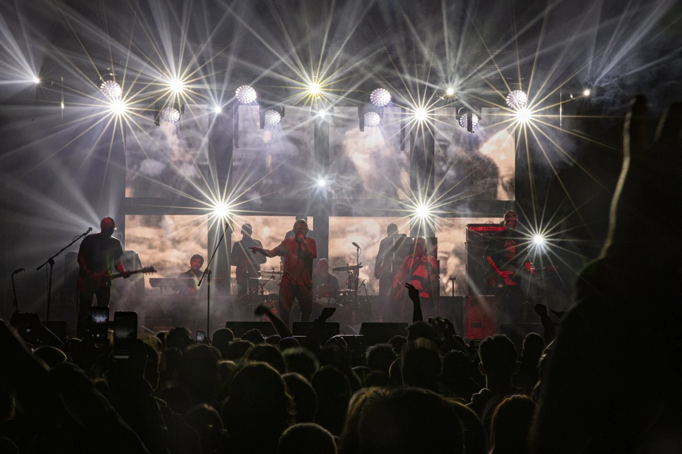 Muzyk grupy Kult stanie przed sądem Tłum ludzi ogląda koncert na żywo, scena oświetlona intensywnymi reflektorami, artyści z instrumentami grają i śpiewają na tle jasnych świateł i dymu scenicznego.