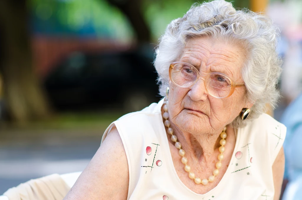 Seniorzy mają często ogromną potrzebę kontroli Starsza kobieta z siwymi włosami, nosząca okulary, koralowy naszyjnik oraz jasną bluzkę siedzi na zewnątrz, patrząc lekko na bok z poważnym wyrazem twarzy.