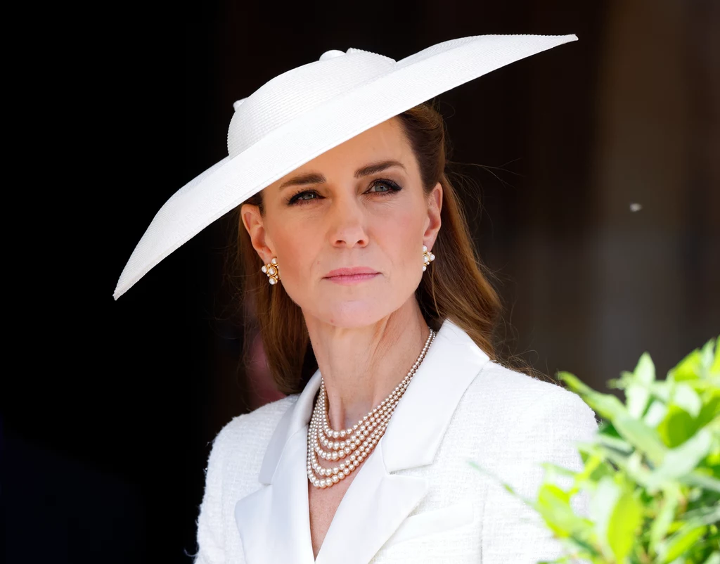 Księżna Kate ostatnio zachwycała swoją formą. Poddani i fani licznie trzymają kciuki za jej powrót do zdrowia Kobieta w eleganckim białym stroju i kapeluszu, z perłową biżuterią, stoi na tle ciemnego tła z zielonymi liśćmi po prawej stronie kadru. Jej wyraz twarzy jest poważny, wzrok skierowany przed siebie.