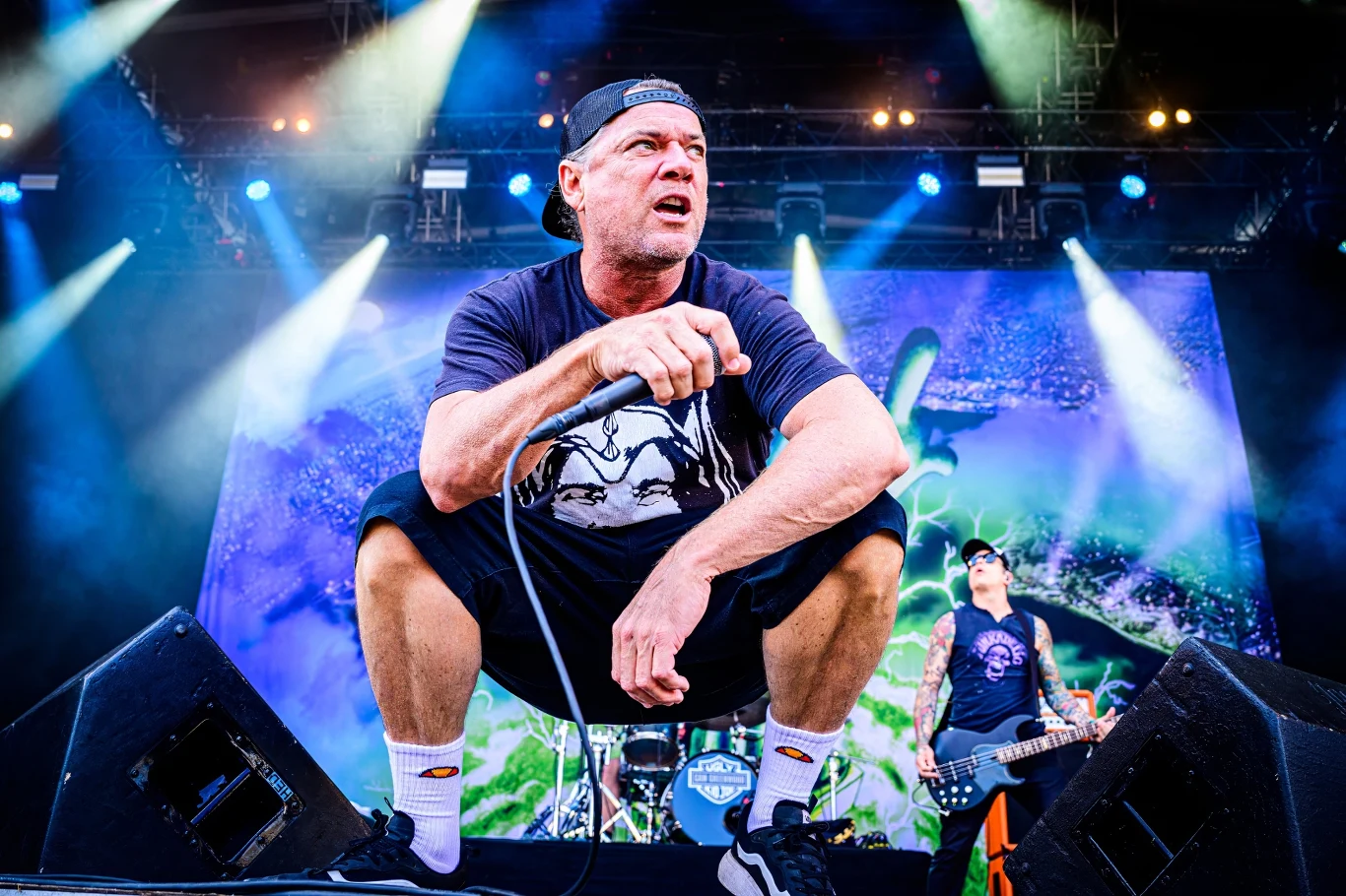 Whitfield Crane stoi na czele Ugly Kid Joe Wokalista zespołu muzycznego w czapce z daszkiem wykonuje utwór na scenie podczas koncertu, za nim widać muzyka grającego na gitarze oraz dynamiczne oświetlenie i kolorową grafikę na ekranie.