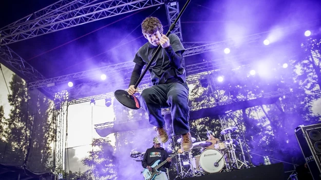 Gitarzysta skaczący w powietrzu na scenie podczas koncertu rockowego, dynamiczne oświetlenie sceniczne w odcieniach fioletu i niebieskiego, widoczny perkusista i gitarzysta basowy, scena na tle drzew i konstrukcji technicznej.
