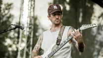 Mężczyzna grający na białej gitarze elektrycznej na scenie plenerowej, ubrany w białą koszulkę bez rękawów i brązową czapkę z daszkiem, otoczony widocznymi elementami oświetlenia i mikrofonem.