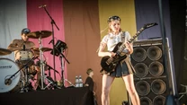 Kobieta grająca na gitarze elektrycznej podczas koncertu na scenie, w tle perkusista, zestaw głośników oraz kolorowa dekoracja sceniczna z pionowymi pasami.