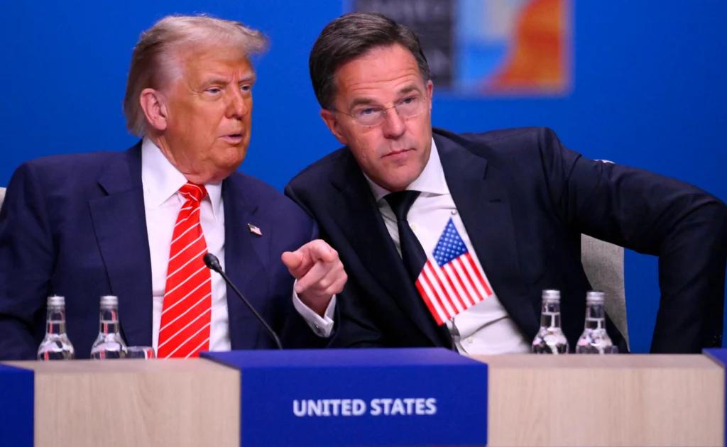 Donald Trump i Mark Rutte podczas szczytu w Hadze Dwóch mężczyzn w garniturach siedzi obok siebie przy stole konferencyjnym, jeden z nich trzyma w ręku miniaturową flagę Stanów Zjednoczonych; przed nimi tabliczka z napisem United States, w tle niebieskie tło i mikrofony.