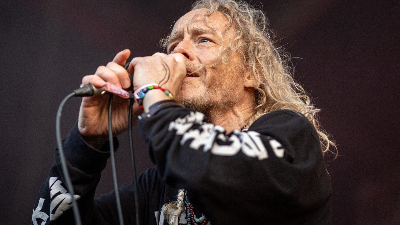 Alan Sparhawk Mężczyzna z długimi, jasnymi włosami śpiewa do mikrofonu podczas występu na scenie, ubrany w czarną bluzę z wyrazistym nadrukiem na rękawach i kolorowe bransoletki na nadgarstku.