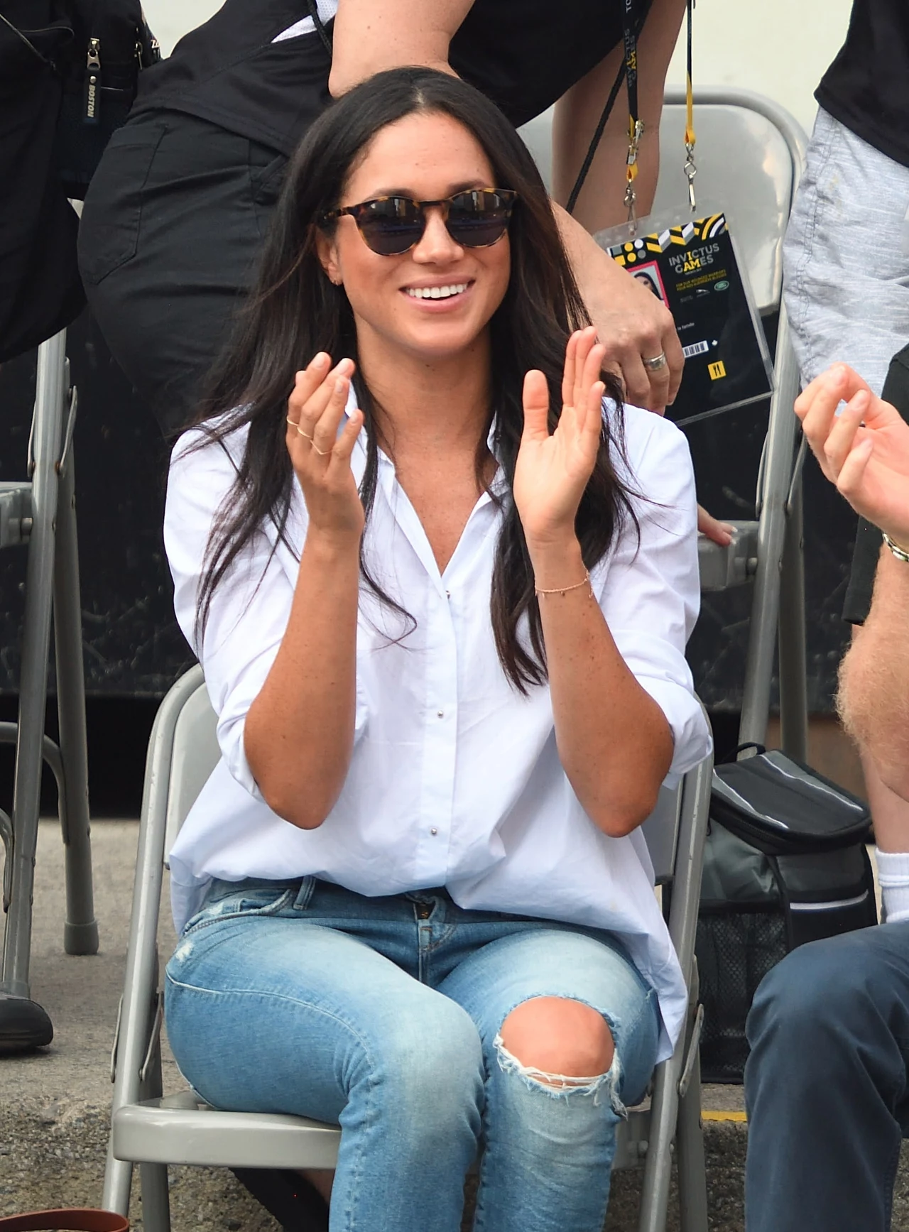 Meghan Markle pierwszy raz publicznie pojawiła się u boku księcia Harry'ego właśnie w klasycznej koszuli Młoda kobieta z długimi ciemnymi włosami ubrana w białą koszulę i niebieskie dżinsy z przetarciami siedzi na krześle, uśmiecha się i klaszcze, mając na sobie ciemne okulary przeciwsłoneczne, wokół znajdują się inne osoby.