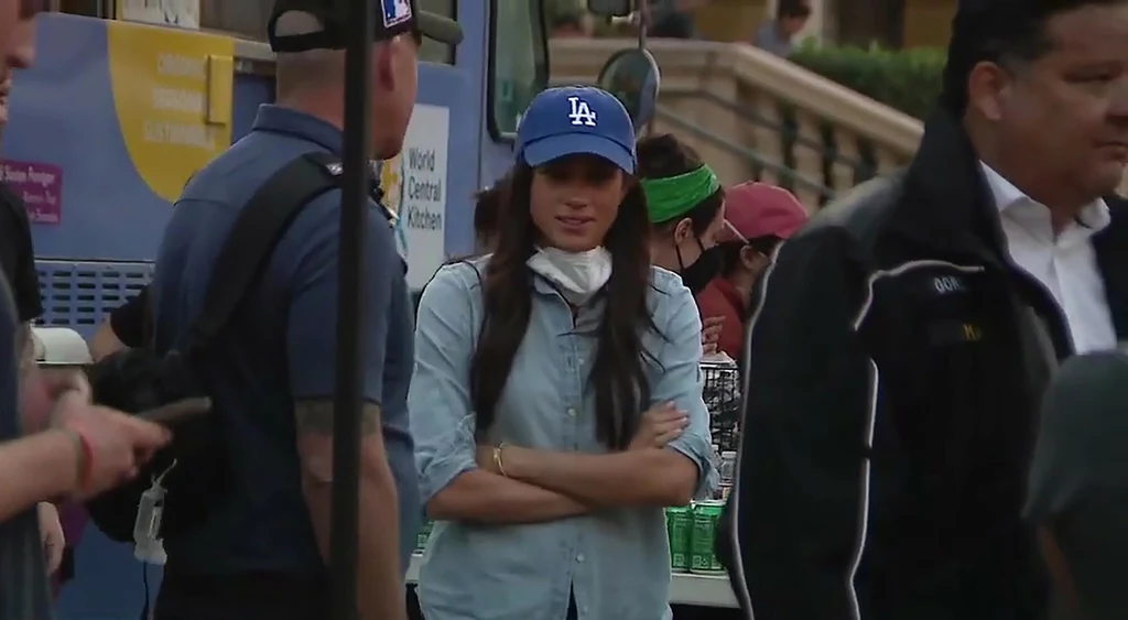 Jeżeli Meghan Markle chce poczuć się jak "dziewczyna z sąsiedztwa" wybiera jeansowe koszule Kobieta w niebieskiej czapce z daszkiem i jasnej koszuli stoi z założonymi rękami wśród kilku osób, w tle punkt wydawania posiłków oraz plastikowe butelki i napoje.