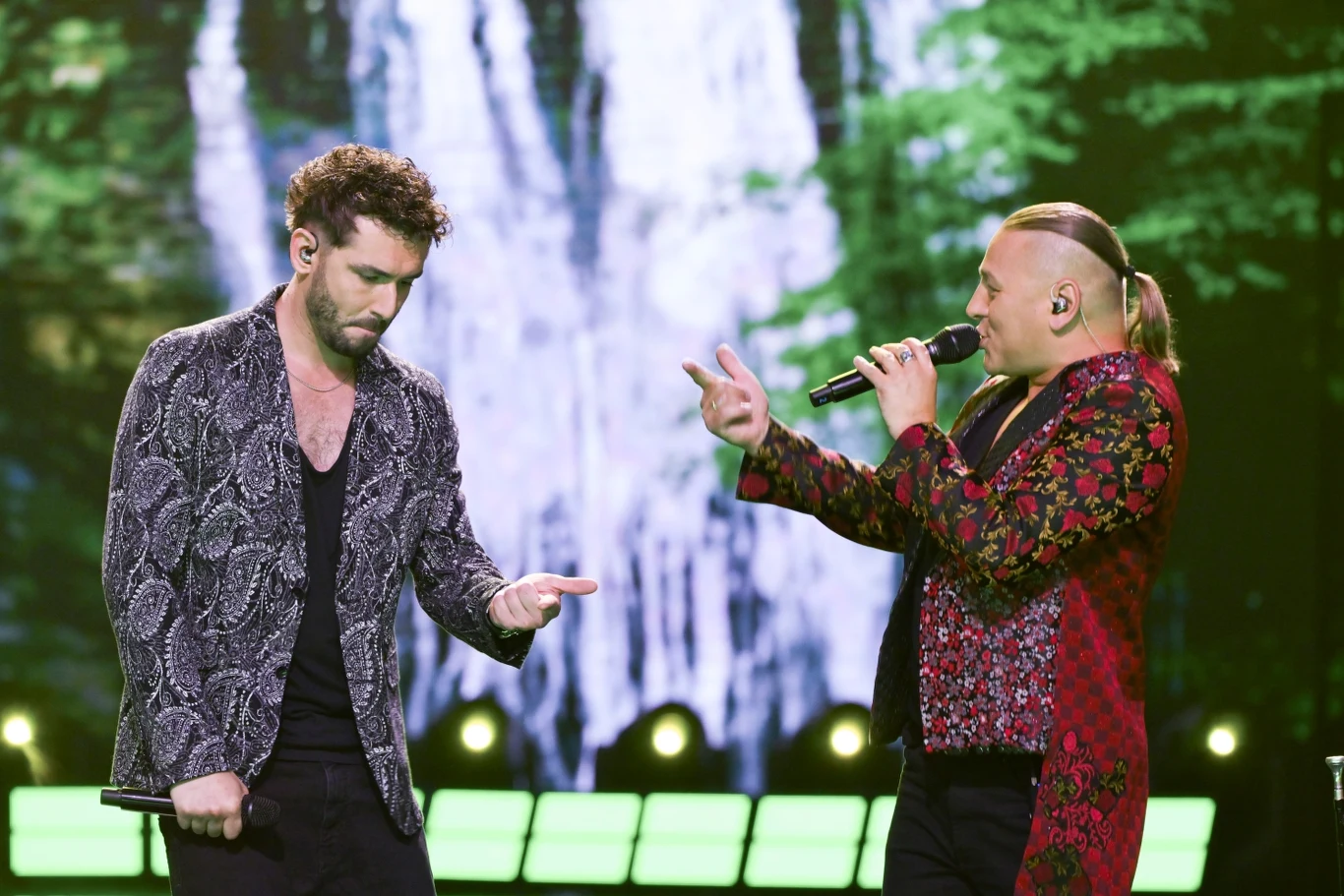 Kamil Bednarek połączył siły z Golec uOrkiestrą Dwóch mężczyzn wykonuje utwór na scenie, jeden śpiewa do mikrofonu, drugi wykonuje gestykulację dłonią, w tle widoczna jest dekoracja przypominająca wodospad i zielone drzewa, scena jest efektownie oświetlona.