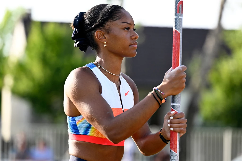 Marie-Julie Bonnin, halowa mistrzyni świata z Nankinu Sportsmenka w stroju lekkoatletycznym trzyma tyczkę do skoku o tyczce, skupiona i przygotowana do konkurencji na tle zieleni i rozmytych budynków.