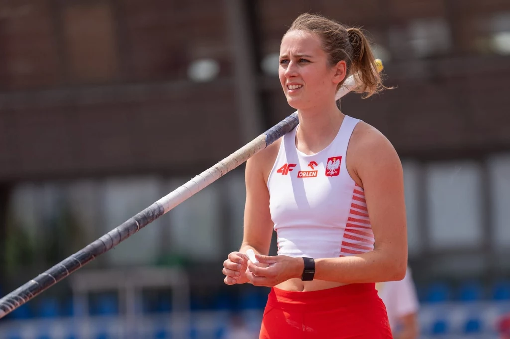 Zofia Gaborska Lekkoatletka w stroju reprezentacji Polski przygotowująca się do skoku o tyczce na stadionie, skupiona na zadaniu, w rękach trzyma tyczkę.