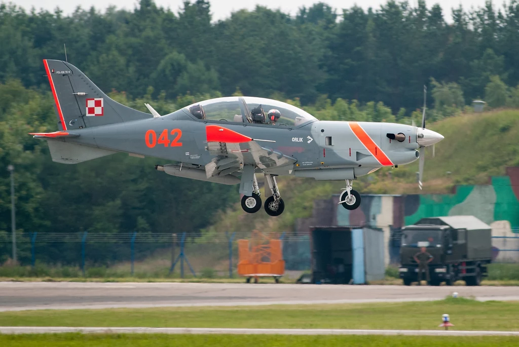 Samolot szkolno-treningowy PZL-130 Orlik Polski wojskowy samolot szkolno-treningowy w szarej kolorystyce z czerwonymi oznaczeniami oraz numerem 042 unosi się nad pasem startowym, w tle widoczne są hangary i zalesiony obszar.