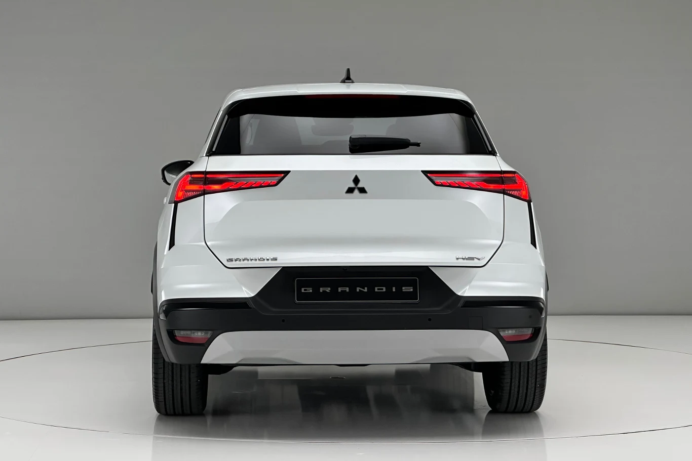Nowoczesne tylne lampy dobrze wkomponowują się w bryłę samochodu. Białe auto marki Mitsubishi Grandis widziane od tyłu, w czystym, dobrze oświetlonym studiu, na tle jednolitej, jasnej ściany i podłogi